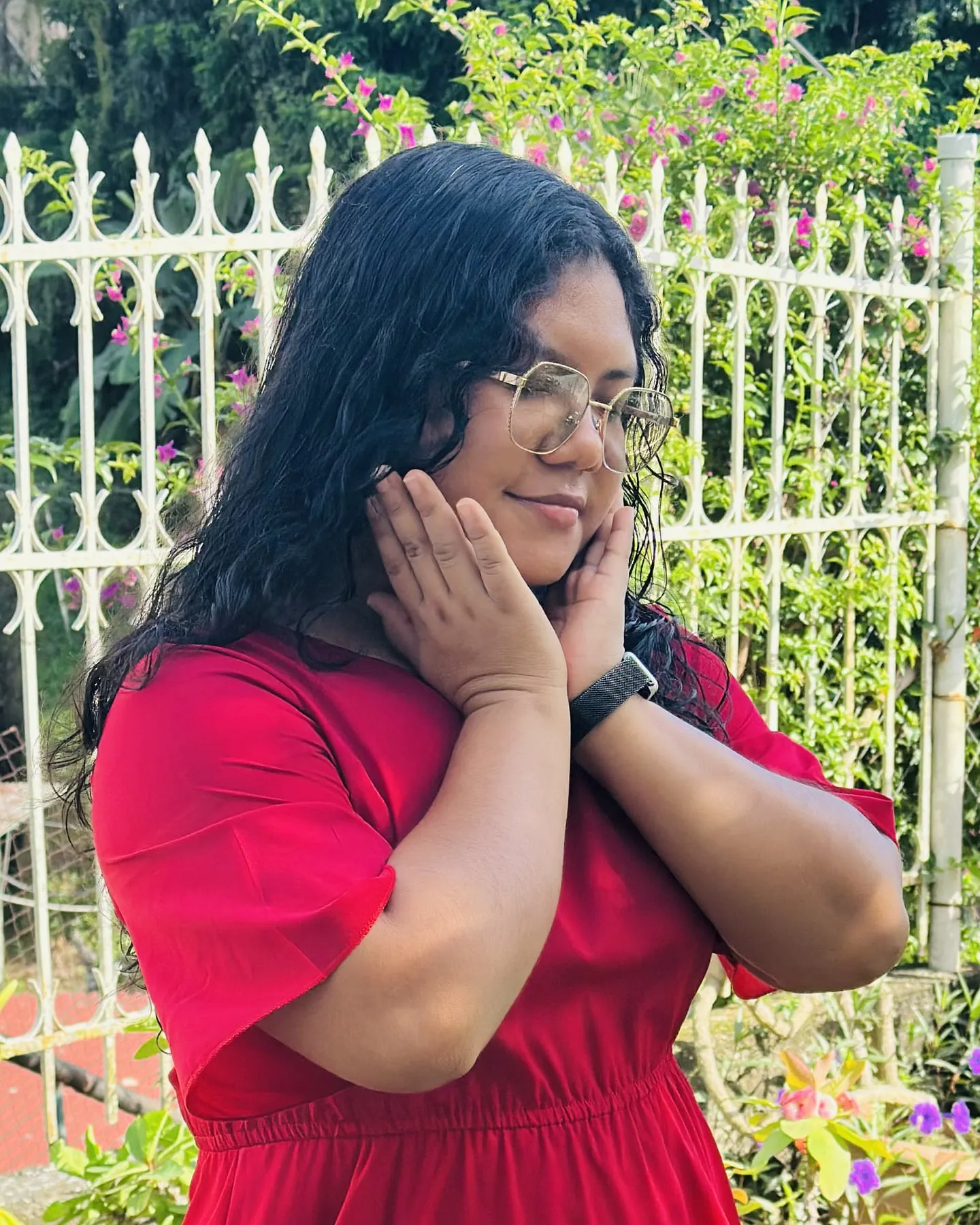 Angie D. Martínez sonriendo, con vestido rojo y lentes, en un jardín.